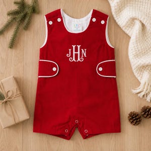 May include: A sleeveless red romper featuring white piping trim, white buttons at the shoulders and sides, and a white embroidered monogram with the letters JHN centered on the chest. The garment includes a white interior label reading LTC 6M.