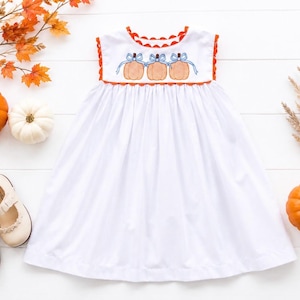 May include: A white sleeveless dress featuring a scalloped orange trim at the neckline and armholes. The bodice displays an embroidered design of three orange pumpkins, each tied with a light blue bow. The skirt is gathered and falls to a hem.