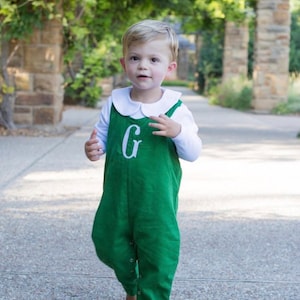 May include: A young child wearing a green overall with a white long-sleeve shirt underneath. The overall has a large white embroidered "G" on the front. The child is walking on a paved path outdoors.