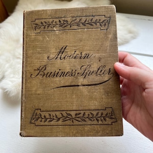 May include: A vintage book titled "Modern Business Speller" with a tan cover. The cover features decorative floral patterns at the top and bottom, with the title in elegant script. The book is held by a hand.