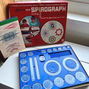 May include: A vintage Spirograph set in a red box with the text "NEW SPIROGRAPH" and illustrations. The blue plastic drawing kit includes various gears, rulers, and drawing tools. The box also includes a booklet with design examples.