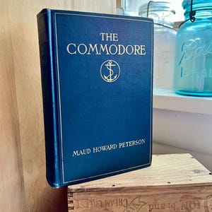 May include: A hardcover book titled "The Commodore" with gold lettering and an anchor emblem on a navy blue cover. The author's name, Maud Howard Peterson, is printed below. The book is displayed on a wooden crate.