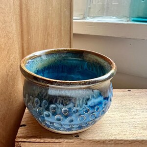 May include: A handmade ceramic bowl with a textured exterior featuring a pattern of raised circles. The bowl has a glossy finish with shades of blue, teal, and brown. The interior is a deep blue, and the rim is a warm brown.