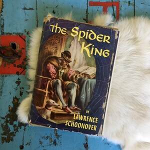 May include: A vintage paperback book titled "The Spider King" by Lawrence Schoonover. The cover features an illustration of a man in a crown and a dog, with a spider web design in the background.