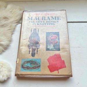 May include: A vintage book titled "Macrame: Creative Design in Knotting" by Dona Z. Meilach. The book cover features four colorful macrame projects, including a hanging planter, a hat, a belt, and a vest.