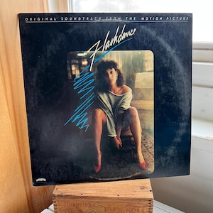 May include: A vintage vinyl record album cover for the movie "Flashdance." The cover is black with the movie title in a stylized font and a photo of a woman in a gray sweater and red heels. The album title is "Original Soundtrack From The Motion Picture."