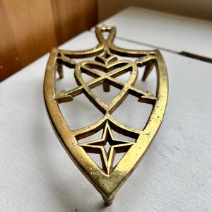 Pode incluir: Um descanso de panela de metal dourado com um design de cora&ccedil;&atilde;o e flechas. O descanso de panela tem a forma de um escudo e tem uma estrela no centro.