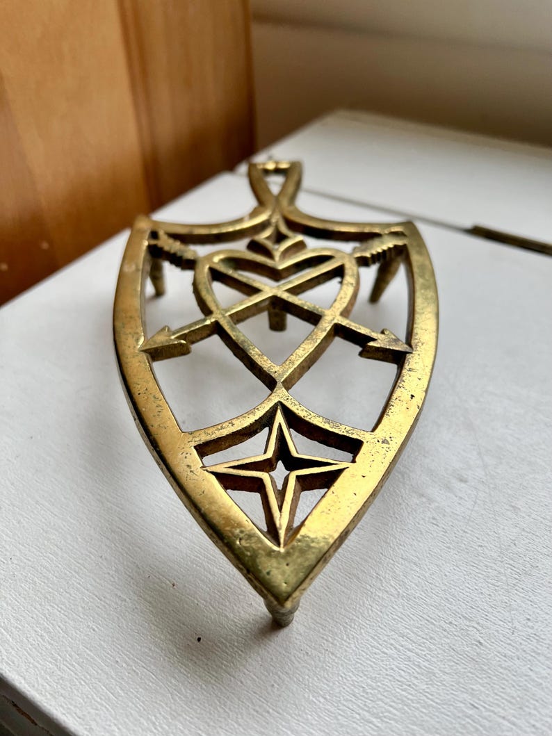 Pode incluir: Um descanso de panela de metal dourado com um design de cora&ccedil;&atilde;o e flechas. O descanso de panela tem a forma de um escudo e tem uma estrela no centro.