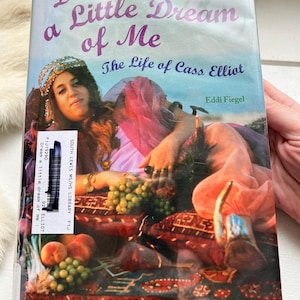 May include: A book titled "Dream a Little Dream of Me" with the subtitle "The Life of Cass Elliot" by Eddi Fiegel. The cover features a photo of a woman in a headscarf, with text in purple and blue. The book is in a library.