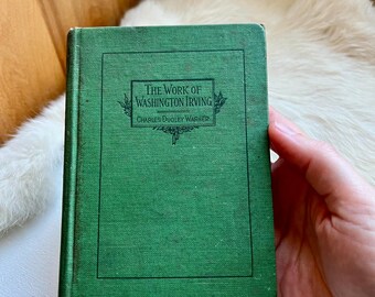 May include: A vintage, hardcover book titled "The Work of Washington Irving" by Charles Dudley Warner. The book has a textured, green cover with the title in white lettering and decorative flourishes. The book is held against a white surface.
