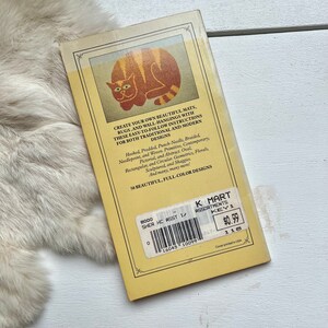 May include: A yellow book with a cat illustration on the cover. The book title is "Create Your Own Beautiful Mats, Rugs, and Wall-Hangings". The book is priced at $0.99 and has a K Mart label.