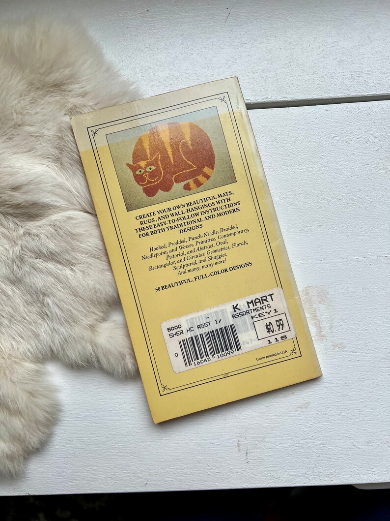 May include: A yellow book with a cat illustration on the cover. The book title is "Create Your Own Beautiful Mats, Rugs, and Wall-Hangings". The book is priced at $0.99 and has a K Mart label.