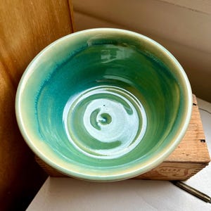May include: A handmade ceramic bowl with a glossy finish. The bowl features a gradient of colors, transitioning from a pale yellow rim to a vibrant turquoise and green interior. The bowl is round and has a smooth, polished surface.
