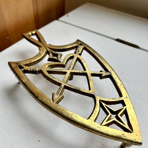 Pode incluir: Um descanso de panela de metal dourado em forma de escudo com um design de cora&ccedil;&atilde;o e flechas. O descanso de panela tem um design decorativo e intrincado.