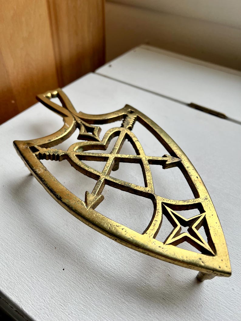 Pode incluir: Um descanso de panela de metal dourado em forma de escudo com um design de cora&ccedil;&atilde;o e flechas. O descanso de panela tem um design decorativo e intrincado.