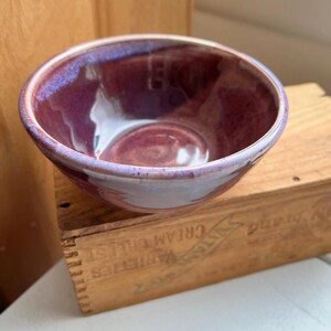 May include: A handmade ceramic bowl with a glossy, deep purple interior and a lighter purple rim. The bowl is round and sits on a wooden crate. The glaze has a subtle, iridescent quality.