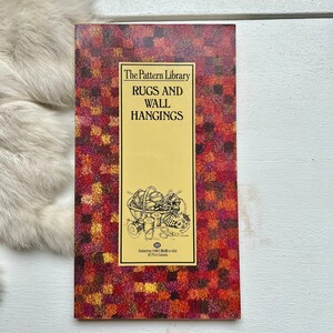 May include: A book titled "The Pattern Library Rugs and Wall Hangings" with a red, orange, and yellow patterned cover. The cover features a black and white illustration of a basket filled with yarn, needles, and scissors.