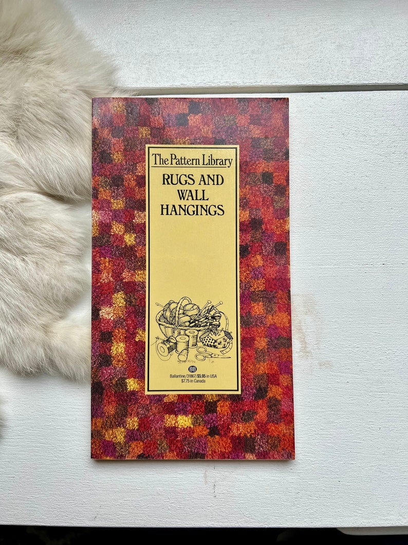 May include: A book titled "The Pattern Library Rugs and Wall Hangings" with a red, orange, and yellow patterned cover. The cover features a black and white illustration of a basket filled with yarn, needles, and scissors.