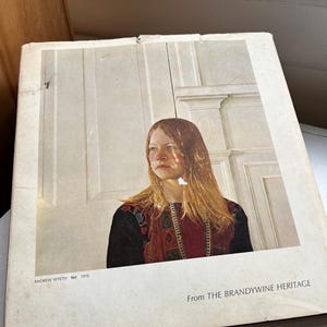 May include: A vintage book cover featuring a portrait of a person with long, light brown hair and a red and black patterned top. The text "From THE BRANDYWINE HERITAGE" is visible, along with the artist's name and the year 1970.