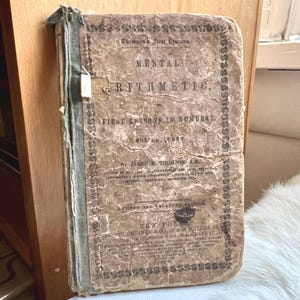 May include: An antique book titled "Mental Arithmetic" with the subtitle "First Lessons in Numbers." The cover is aged with visible wear and tear, featuring faded text and a decorative border. The book is a revised and enlarged edition.