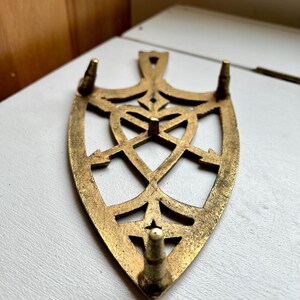 Pode incluir: Um descanso de ferro de metal dourado com um design decorativo e intrincado. O descanso tem tr&ecirc;s pernas pontiagudas.