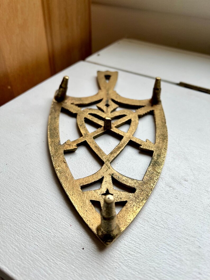 Pode incluir: Um descanso de ferro de metal dourado com um design decorativo e intrincado. O descanso tem tr&ecirc;s pernas pontiagudas.