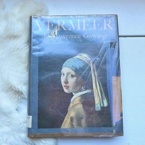 May include: A vintage hardcover book titled "Vermeer" by Lawrence Gowing. The cover features a painting of a woman with a blue headscarf and a pearl earring.