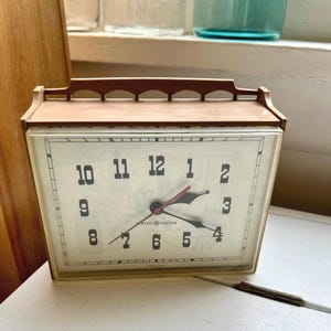 May include: Vintage General Electric clock with a cream-colored face and black numerals. The clock has a brown frame and red second hand. The clock face has a decorative design and the words "General Electric" are printed on the face.