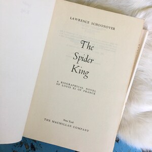May include: A white book with the title "The Spider King" by Lawrence Schoonover. The book is a biographical novel about Louis XI of France and was published by The Macmillan Company in New York.