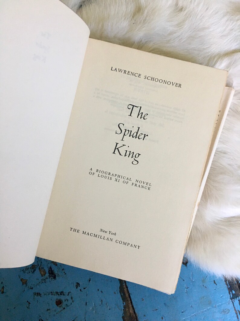 May include: A white book with the title "The Spider King" by Lawrence Schoonover. The book is a biographical novel about Louis XI of France and was published by The Macmillan Company in New York.