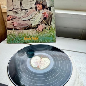 May include: A vintage vinyl record and album cover featuring James Taylor. The album cover shows a man in a brown suit, reclining on grass in front of a stone wall. The record is black with a white label.