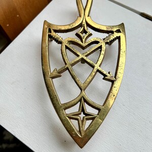 Pode incluir: Um descanso de panela de metal dourado em forma de cora&ccedil;&atilde;o com flechas que o atravessam. O descanso de panela tem um design decorativo com uma estrela na parte inferior.