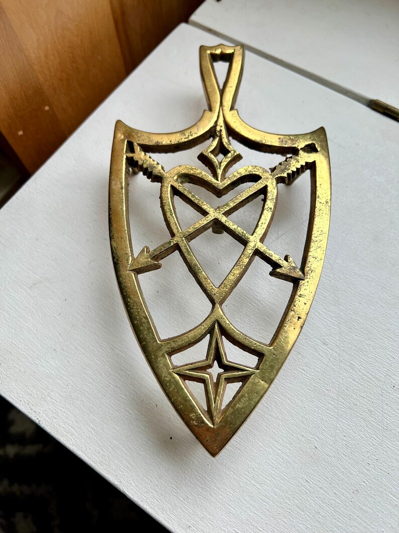 Pode incluir: Um descanso de panela de metal dourado em forma de cora&ccedil;&atilde;o com flechas que o atravessam. O descanso de panela tem um design decorativo com uma estrela na parte inferior.