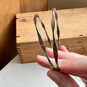 May include: A set of three silver-toned, twisted metal bangles. The bangles have a smooth, reflective surface and a unique, wavy design. The jewelry is held in a hand, with a wooden box in the background.