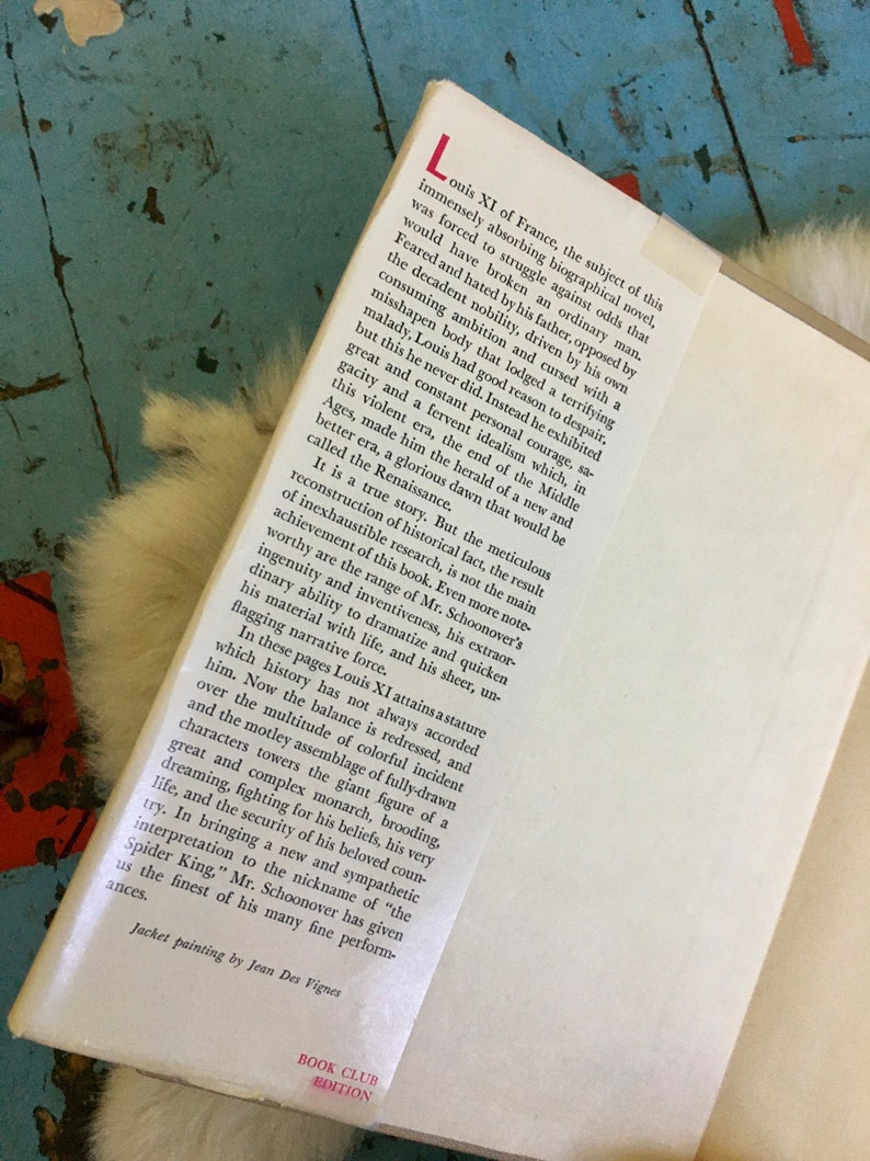 May include: A book cover with a red and white book club edition label. The cover features a painting of a man in a dark blue and gold robe with a white collar. The text on the cover reads "Louis XI of France, the subject of this immensely absorbing biographical novel..."