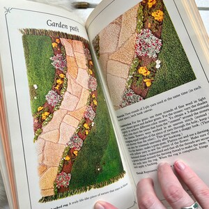 May include: A latch hook rug depicting a garden path with green grass, brown stones, yellow flowers, and pink flowers. The rug is a close-up of the bottom right corner of the rug, showing the fringe and the text "Latch hooked rug A truly life-like piece of nature that stays in bloom all year long."