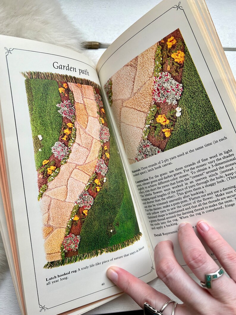 May include: A latch hook rug depicting a garden path with green grass, brown stones, yellow flowers, and pink flowers. The rug is a close-up of the bottom right corner of the rug, showing the fringe and the text "Latch hooked rug A truly life-like piece of nature that stays in bloom all year long."