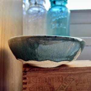 May include: A shallow ceramic bowl with a speckled cream base and a dark teal glazed rim. The bowl sits atop a wooden crate with the text "VITAMIN D" and "CREAM CHEESE" visible. Two glass jars are positioned in the background behind the bowl.