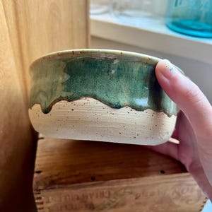 May include: A ceramic bowl featuring a speckled cream-colored base and a glossy, dark green glaze that drips down from the rim. The bowl has a smooth, rounded shape with a distinct contrast between the textured, unglazed bottom and the shiny top.