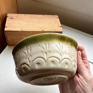 May include: A cream-colored ceramic bowl with a green rim and embossed floral design. The bowl is held in a hand and has a rounded shape with a decorative pattern around the sides. A wooden box is in the background.