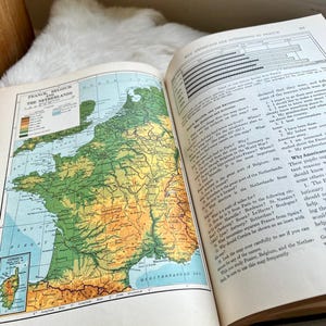 May include: Open vintage book with a detailed map of France, Belgium, and the Netherlands. The map features various colors to indicate land elevation and geographical features. Text on the right side discusses American interest in France.
