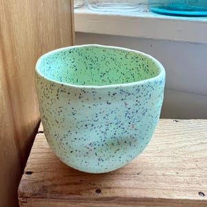 May include: A light green ceramic cup with an irregular, hand-formed shape and a speckled finish featuring small dark blue, brown, and orange dots scattered across the exterior surface. The cup has a wide, open top and a rounded base.
