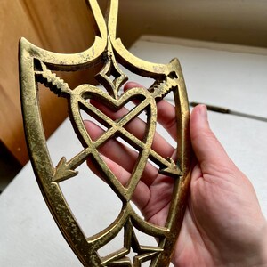 Pode incluir: Um descanso de panela de metal dourado em forma de cora&ccedil;&atilde;o com flechas que o atravessam. O descanso de panela tem um design decorativo com uma estrela na parte inferior.