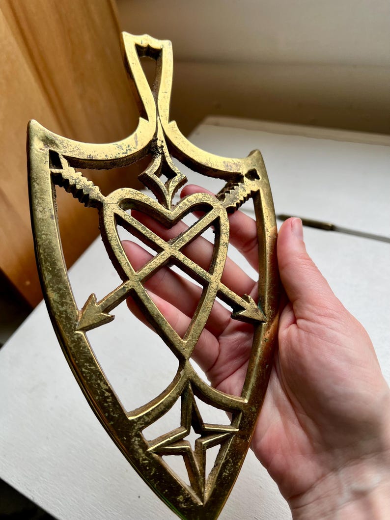 Pode incluir: Um descanso de panela de metal dourado em forma de cora&ccedil;&atilde;o com flechas que o atravessam. O descanso de panela tem um design decorativo com uma estrela na parte inferior.