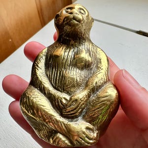 May include: A small, solid brass monkey figurine sitting with its legs crossed and looking up. The monkey is detailed with textured fur and has a realistic appearance.
