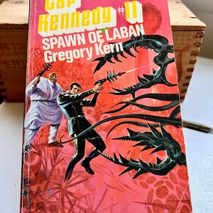 May include: A vintage paperback science fiction book titled Cap Kennedy #11 Spawn of Laban by Gregory Kern. The cover features a bright red and pink background with a dark-skinned figure in white robes and a man in a black uniform firing a ray gun.
