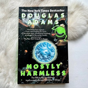 May include: A book cover with a black background and a green alien with a big smile. The title of the book is "Mostly Harmless" and the author is Douglas Adams. The book is the fifth in the Hitchhiker's Trilogy.