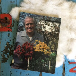 Puede incluir: Un libro vintage titulado "Crockett's Indoor Garden" que muestra a un hombre sosteniendo un ramo de flores rojas y amarillas. La portada del libro tiene un fondo blanco con texto negro.