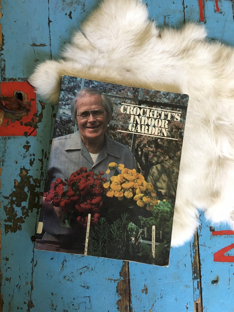 Puede incluir: Un libro vintage titulado "Crockett's Indoor Garden" que muestra a un hombre sosteniendo un ramo de flores rojas y amarillas. La portada del libro tiene un fondo blanco con texto negro.