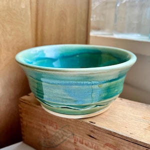 May include: A handmade ceramic bowl with a turquoise and teal glaze. The bowl has a rounded shape with a slightly flared rim and sits on a wooden crate. The interior of the bowl reflects the light, showcasing the depth of the glaze.